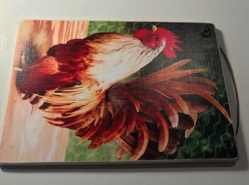 May include: Rectangular wooden cutting board featuring a detailed image of a rooster. The rooster has red, brown, and white feathers, with a red comb and wattle. A leather loop is attached for hanging. The background includes a chicken wire fence.