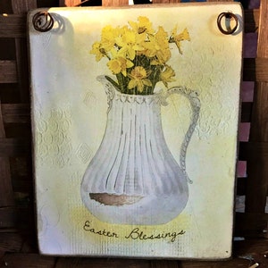 Può includere: Targa rettangolare in legno con una brocca bianca piena di narcisi gialli. La brocca è posta su uno sfondo giallo pallido con un motivo decorativo. Le parole "Easter Blessings" sono scritte in corsivo. La targa ha un gancio di filo.