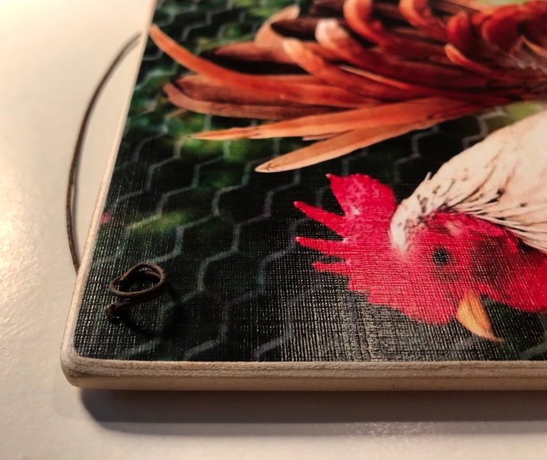 May include: A rectangular wooden sign featuring a rooster illustration. The rooster has red, white, and brown feathers, set against a green mesh background. A dark brown string is attached for hanging.