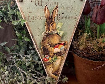 Può includere: Un cartello di legno triangolare con uno sfondo verde invecchiato che presenta un coniglio marrone che tiene un cesto di uova di Pasqua colorate. Il cartello recita "Wishing you every Easter Blessing".