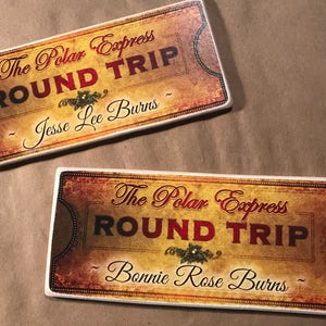 May include: Two vintage-style Polar Express train tickets with the text "The Polar Express Round Trip" in red and gold lettering. The top ticket has the name "Jesse Lee Burns" and the bottom ticket has the name "Bonnie Rose Burns".