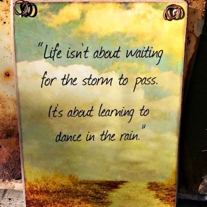 May include: A rustic wooden sign with a painted landscape background and a quote about learning to dance in the rain. The quote reads, "Life isn't about waiting for the storm to pass. It's about learning to dance in the rain."
