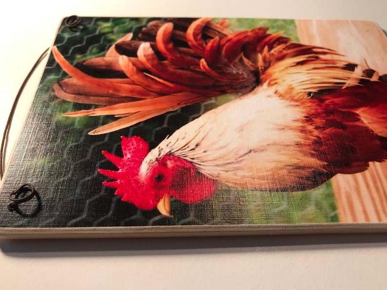 May include: A wooden cutting board featuring a detailed image of a rooster. The rooster has vibrant red, brown, and white feathers, with a red comb and wattle. The background includes a wire fence and a wooden surface.