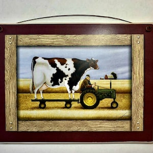 May include: A framed wood sign with a painting of a farmer driving a green tractor with a black and white cow on a wooden platform behind him. The background is a field of hay.