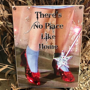Può includere: Un cartello di legno con un'immagine di Dorothy de Il Mago di Oz che indossa scarpe di rubino rosso. Il cartello dice "Non c'è posto come casa".