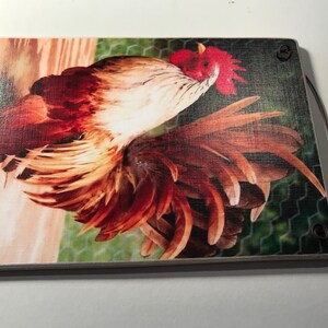 May include: A rectangular wooden cutting board featuring a vibrant rooster illustration. The rooster has red, brown, and white feathers, with a red comb. The background includes a wooden fence and chicken wire. A brown handle is attached.