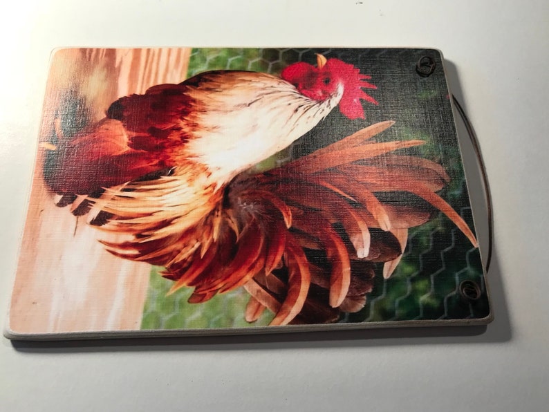May include: A rectangular wooden cutting board featuring a vibrant rooster illustration. The rooster has red, brown, and white feathers, with a red comb. The background includes a wooden fence and chicken wire. A brown handle is attached.