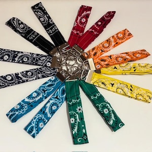 Può includere: Una collezione di dieci portachiavi da polso in tessuto disposti in un motivo circolare. Ogni cinturino presenta una distinta stampa paisley bandana in colori che includono nero, blu navy, verde acqua, verde, giallo, arancione e rosso, rifiniti con componenti in metallo argentato.
