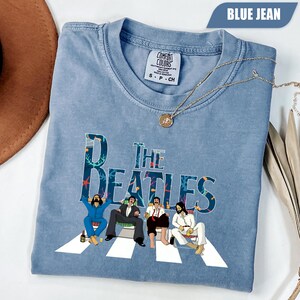 Puede incluir: Una camiseta color azul jean con un gráfico de The Beatles sentados en sillas sobre un camino de rayas blancas. El gráfico incluye patrones de arrecifes de coral dentro de las letras. Un collar dorado descansa sobre el cuello de la camiseta.