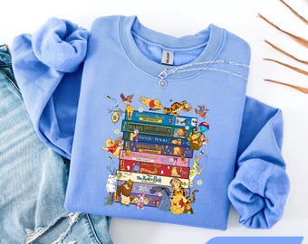 Könnte beinhalten: Ein Carolina-blaues Sweatshirt mit Rundhalsausschnitt und einem Grafikdruck eines Stapels bunter Bücher mit Winnie Puuh-Geschichten. Das Design enthält Illustrationen von Puuh, Tigger, Ferkel, I-Aah und anderen Charakteren um den Bücherstapel herum.