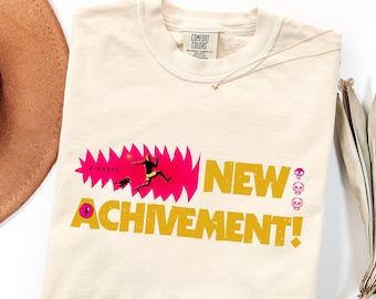 Pode incluir: Uma t-shirt cor de marfim com um gráfico que inclui o texto "NEW ACHIVEMENT!" em amarelo, uma forma dentada cor-de-rosa, uma pequena figura a correr e três ícones de caveiras cor-de-rosa. A etiqueta Comfort Colors está no colarinho.