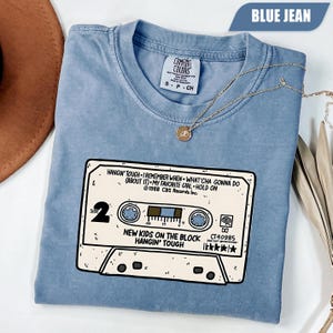 Puede incluir: Una camiseta color azul jean con un gráfico de un casete etiquetado como New Kids on the Block Hangin' Tough. El estampado incluye títulos de canciones, el año 1988 y el texto CBS Records Inc. La prenda tiene cuello redondo y una etiqueta.