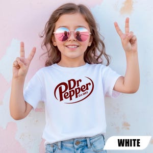 Puede incluir: Una camiseta blanca de manga corta con el logotipo de Dr Pepper en rojo oscuro y el texto Est.1885 impreso en el pecho. La camiseta se combina con unos vaqueros azul claro y el dise&ntilde;o est&aacute; centrado en la parte frontal de la prenda.