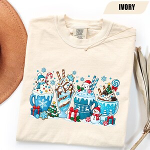 May include: An ivory t-shirt with a graphic print featuring four festive mugs and glasses filled with holiday treats, including snowmen, candy canes, Christmas trees, wrapped gifts, and snowflakes. The shirt has a Comfort Colors brand label.