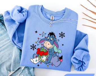 Könnte beinhalten: Ein Carolina-blaues Sweatshirt mit Rundhalsausschnitt und einer Grafik von I-Aah, der sich in bunten Weihnachtsbeleuchtung verheddert hat. Die Figur hält ein grünes Geschenk mit roter Schleife, umgeben von schwarzen Schneeflockensymbolen.