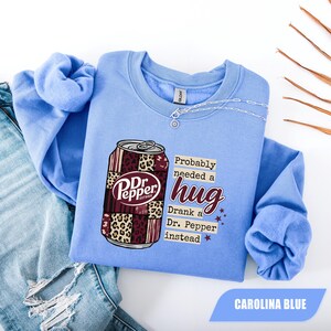 May include: A Carolina blue crewneck sweatshirt featuring a graphic of a soda can with a patchwork design of maroon and leopard print patterns. The text reads: Probably needed a hug, Drank a Dr. Pepper instead, with small maroon stars.