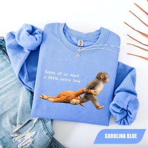 Puede incluir: Una sudadera de cuello redondo color azul Carolina con un gráfico de un mono gris arrastrando a un mono de peluche marrón por el suelo. El texto "Some of us need a little extra love" está impreso en blanco sobre los monos. La sudadera tiene mangas largas.