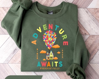 Könnte beinhalten: Ein olivgrüner Rundhals-Pullover mit einer bunten Grafik eines Hauses, das von einem großen Ballonbündel in die Luft gehoben wird. Der Text lautet ADVENTURE AWAITS, SINCE 2009 und PARADISE FALLS in bunten Farben, umgeben von orangefarbenen Wolken.