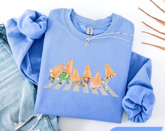 Op de afbeelding: Een Carolina blauwe sweater met ronde hals en een grafische print van vijf geanimeerde figuren die over een zebrapad lopen, elk met een oranje verkeerskegel op hun hoofd. De sweater heeft lange mouwen en een geribbelde kraag.