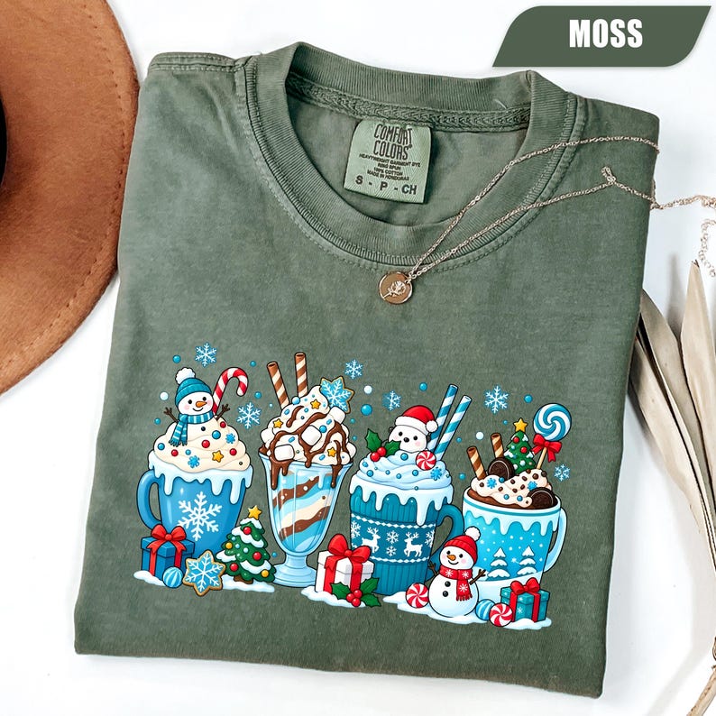May include: A moss green crewneck t-shirt featuring a graphic print of four mugs filled with hot cocoa, topped with whipped cream, candy canes, and holiday decorations. The shirt has a tag labeled Comfort Colors and a delicate gold necklace.