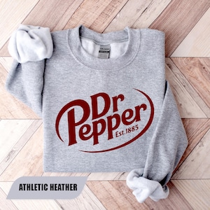 May include: A light gray crewneck sweatshirt featuring the dark red Dr Pepper logo and the text Est. 1885 printed on the front. The garment has long sleeves with ribbed cuffs and a Gildan brand label inside the collar. The color is athletic heather.