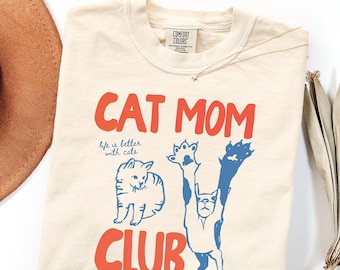 Op de afbeelding: Een ivoorkleurig t-shirt met de rode tekst CAT MOM CLUB en de blauwe tekst life is better with cats. De afbeelding bevat twee blauwe kattenillustraties, één zittend en één uitrekkend met de pootjes omhoog. Het shirt heeft een Comfort Colors label.