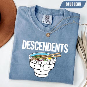 Puede incluir: Una camiseta color azul jean con cuello redondo y etiqueta de la marca Comfort Colors. La prenda presenta el texto blanco DESCENDENTS sobre un gráfico de un tazón de ramen con gafas y palillos descansando sobre la parte superior del tazón.