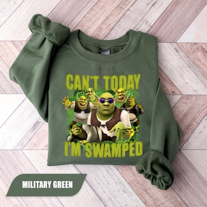 May include: A military green crewneck sweatshirt featuring a graphic print of multiple Shrek characters. The text CAN'T TODAY I'M SWAMPED is printed in yellow above and below the characters. The central figure wears dark sunglasses.