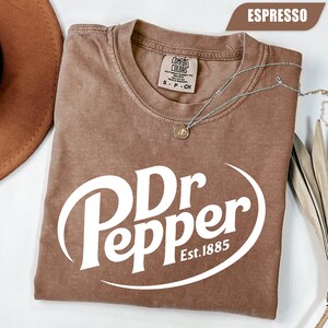 Puede incluir: Una camiseta marr&oacute;n de manga corta con cuello redondo y un estampado gr&aacute;fico blanco que dice Dr Pepper Est. 1885. La camiseta presenta una etiqueta de la marca Comfort Colors en el cuello interior y se muestra con un collar de plata encima.