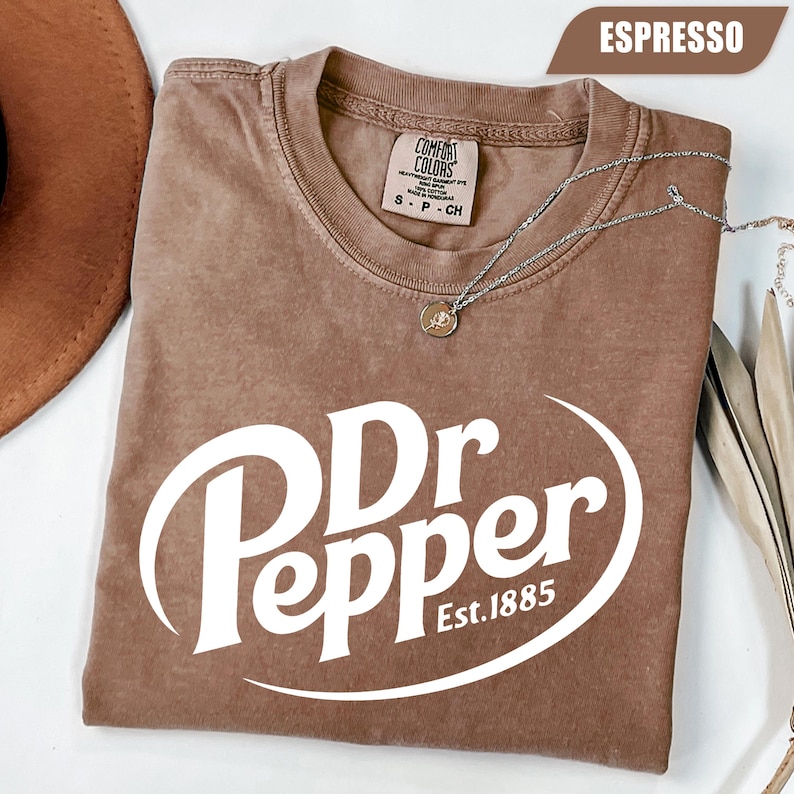Puede incluir: Una camiseta marr&oacute;n de manga corta con cuello redondo y un estampado gr&aacute;fico blanco que dice Dr Pepper Est. 1885. La camiseta presenta una etiqueta de la marca Comfort Colors en el cuello interior y se muestra con un collar de plata encima.