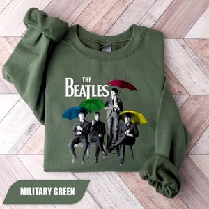 Könnte beinhalten: Ein militärgrüner Rundhals-Pullover mit einem grafischen Aufdruck der Beatles. Das Design zeigt die vier Bandmitglieder in Anzügen, die jeweils einen bunten Regenschirm in Rot, Grün, Blau oder Gelb halten, mit dem Text The Beatles darüber.