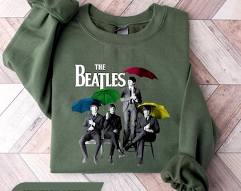 Op de afbeelding: Een militair groene sweater met ronde hals en een grafische print van The Beatles. Het ontwerp toont de vier bandleden in pak, elk met een kleurrijke paraplu in rood, groen, blauw of geel, met de tekst The Beatles erboven afgebeeld.