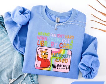 Könnte beinhalten: Ein hellblaues Sweatshirt mit Rundhalsausschnitt und einer Grafik der Figur Arthur Read sowie dem Text "Having fun isn't hard when you've got a library card" über einer bunten Bibliotheksausweis-Illustration mit Barcode und der Nummer 2101908.