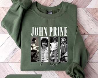 May include: A military green crewneck sweatshirt featuring the text JOHN PRINE in white capital letters above four vertical black and white photographic panels of the musician. A black signature is printed below the photo panels on the front.