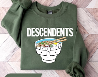 Op de afbeelding: Een militair groene sweater met ronde hals en de witte tekst DESCENDENTS boven een afbeelding van een witte ramenkom met een gezicht met een zwart montuur, gevuld met noedels, groenten en een ei, met twee houten eetstokjes.