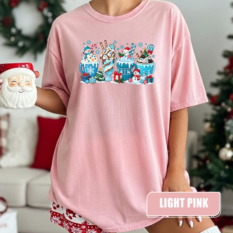 May include: A light pink short-sleeve t-shirt featuring a graphic print of four blue mugs filled with hot cocoa, whipped cream, candy canes, and small snowmen, surrounded by snowflakes, Christmas trees, and wrapped gift boxes.