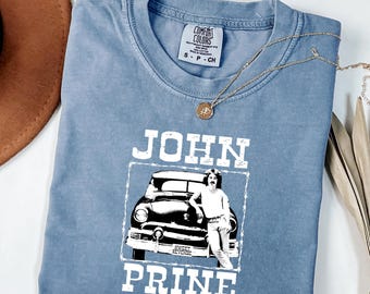 Könnte beinhalten: Ein blaues T-Shirt in Jeansoptik mit einem weißen Grafikdruck von John Prine, der an einem Oldtimer lehnt. Der Text "JOHN PRINE" ist in großen weißen Blockbuchstaben über und unter dem Bild gedruckt, mit "SWEET REVENGE" auf dem Nummernschild.