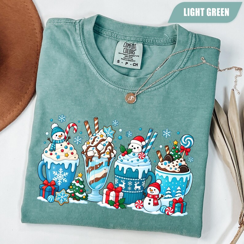 May include: A light green short-sleeve t-shirt featuring a graphic print of four festive mugs and glasses filled with holiday treats, including snowmen, candy canes, Christmas trees, and wrapped gifts, with the Comfort Colors brand label visible.