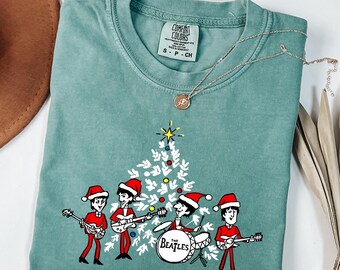 Könnte beinhalten: Ein hellgrünes T-Shirt mit einer Grafik von vier Zeichentrickfiguren in roten Anzügen und Weihnachtsmützen, die um einen stilisierten weißen Weihnachtsbaum herum Musikinstrumente spielen. Das T-Shirt hat ein Comfort Colors Etikett am Kragen.