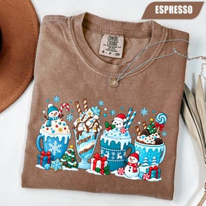 May include: An espresso-colored t-shirt featuring a graphic print of four festive mugs filled with hot cocoa, whipped cream, and holiday treats like candy canes, small Christmas trees, gift boxes, and snowmen, with snowflakes scattered throughout.