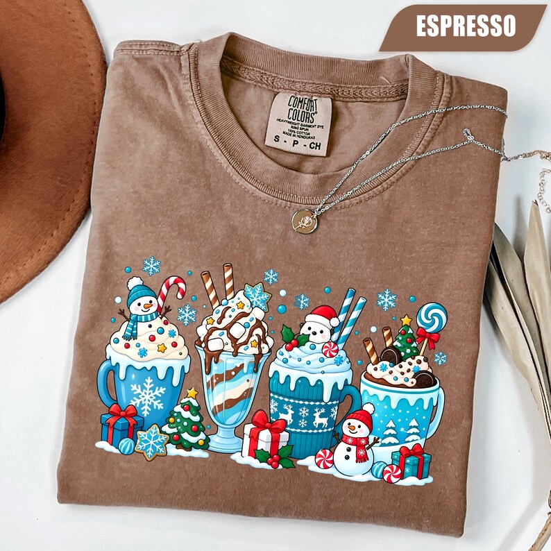 May include: An espresso-colored t-shirt featuring a graphic print of four festive mugs filled with hot cocoa, whipped cream, and holiday treats like candy canes, small Christmas trees, gift boxes, and snowmen, with snowflakes scattered throughout.