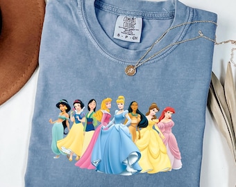Könnte beinhalten: Ein blaues T-Shirt in Jeansoptik mit einem Grafikdruck von acht Disney-Prinzessinnen, die in einer Reihe stehen. Das Shirt hat einen Rundhalsausschnitt und ein Comfort Colors Markenetikett am Innenkragen. Eine goldfarbene Kette liegt auf dem Stoff.