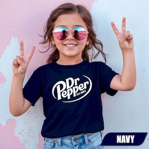 Puede incluir: Una camiseta azul marino de manga corta con el logotipo blanco de Dr Pepper y el texto Est.1885 impreso en el pecho. La prenda se combina con unos vaqueros de mezclilla azul claro y gafas de sol redondas con lentes reflectantes rosas.