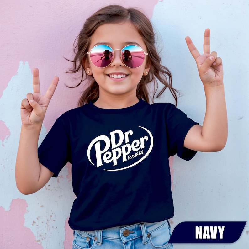 Puede incluir: Una camiseta azul marino de manga corta con el logotipo blanco de Dr Pepper y el texto Est.1885 impreso en el pecho. La prenda se combina con unos vaqueros de mezclilla azul claro y gafas de sol redondas con lentes reflectantes rosas.