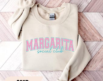 Op de afbeelding: Een zandkleurige sweater met ronde hals voorzien van de tekst MARGARITA in een roze en blauw collegiaal lettertype, met daaronder de woorden social club in een blauw cursief handschrift. Het kledingstuk heeft lange mouwen en geribbelde boorden.
