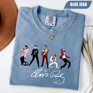 May include: A blue jean colored short-sleeve t-shirt featuring five graphic illustrations of Elvis Presley in various poses and outfits, with the signature Elvis Presley written in white script below the figures and a Comfort Colors brand tag.