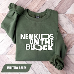 Puede incluir: Una sudadera de cuello redondo y manga larga en color verde militar con el texto blanco NEW KIDS ON THE BLOCK impreso en una fuente negrita sin remates en el pecho. La prenda presenta puños y cuello acanalados de diseño sólido.