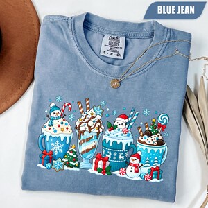 May include: A blue jean colored t-shirt featuring a graphic print of four mugs filled with hot cocoa and milkshakes, topped with whipped cream, candy canes, and marshmallows, surrounded by snowmen, Christmas trees, and gift boxes.