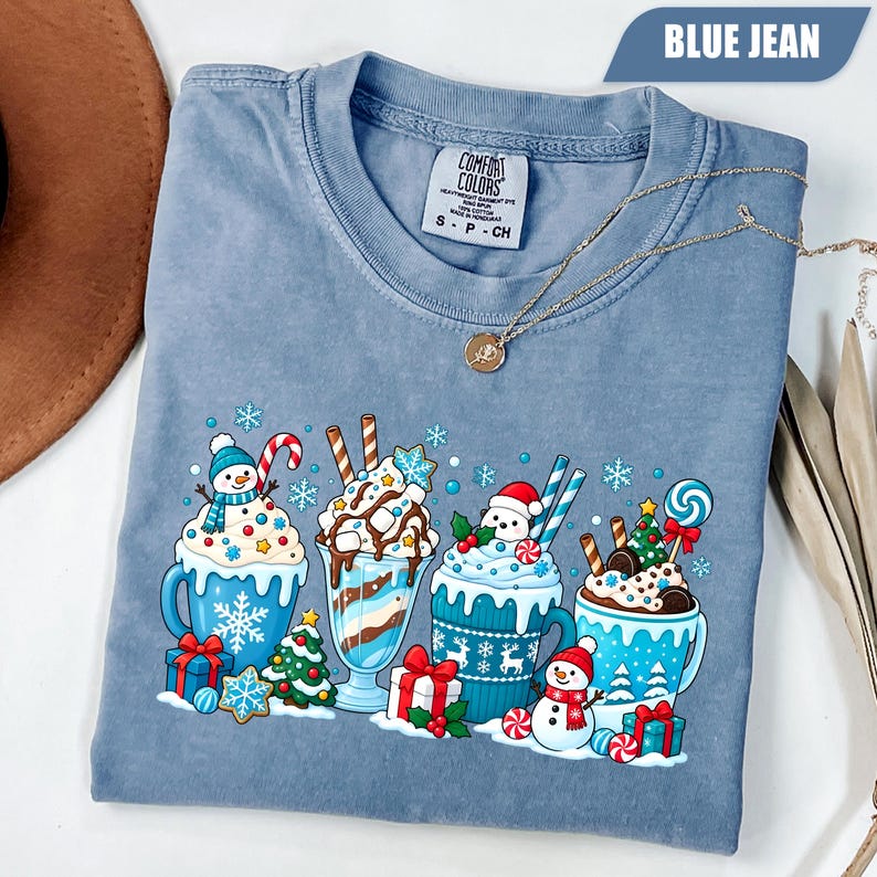 May include: A blue jean colored t-shirt featuring a graphic print of four mugs filled with hot cocoa and milkshakes, topped with whipped cream, candy canes, and marshmallows, surrounded by snowmen, Christmas trees, and gift boxes.
