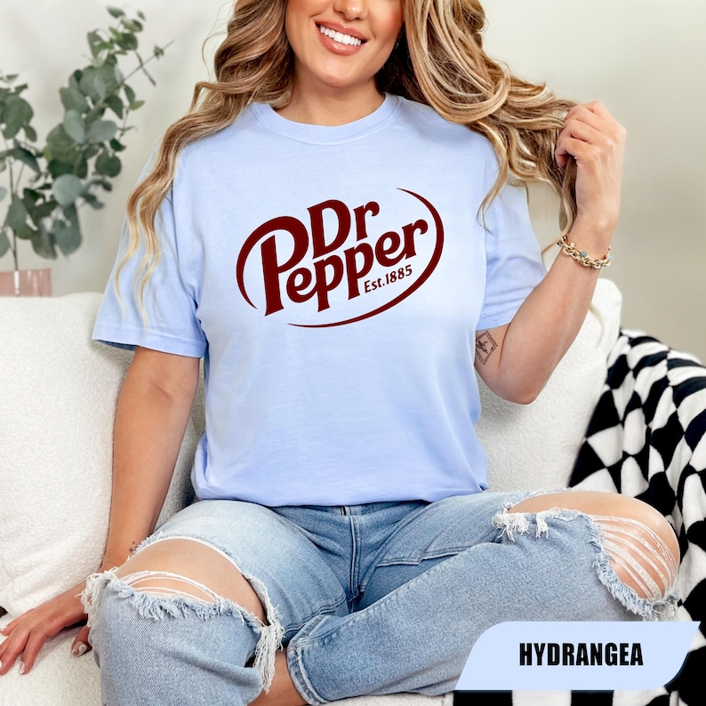 Puede incluir: Una camiseta de manga corta de color azul claro con el logotipo de Dr Pepper en rojo oscuro y el texto Est.1885 impreso en el centro. La prenda tiene cuello redondo y un corte holgado, mostrando el nombre de la marca en una fuente cursiva.